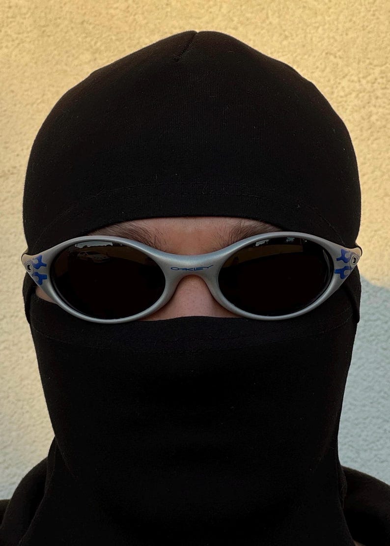 Puede incluir: Gafas de sol con montura plateada, lentes oscuras y detalles azules. Las gafas se llevan con un pasamonta&ntilde;as negro. La imagen es un primer plano que se centra en las gafas y el pasamonta&ntilde;as.