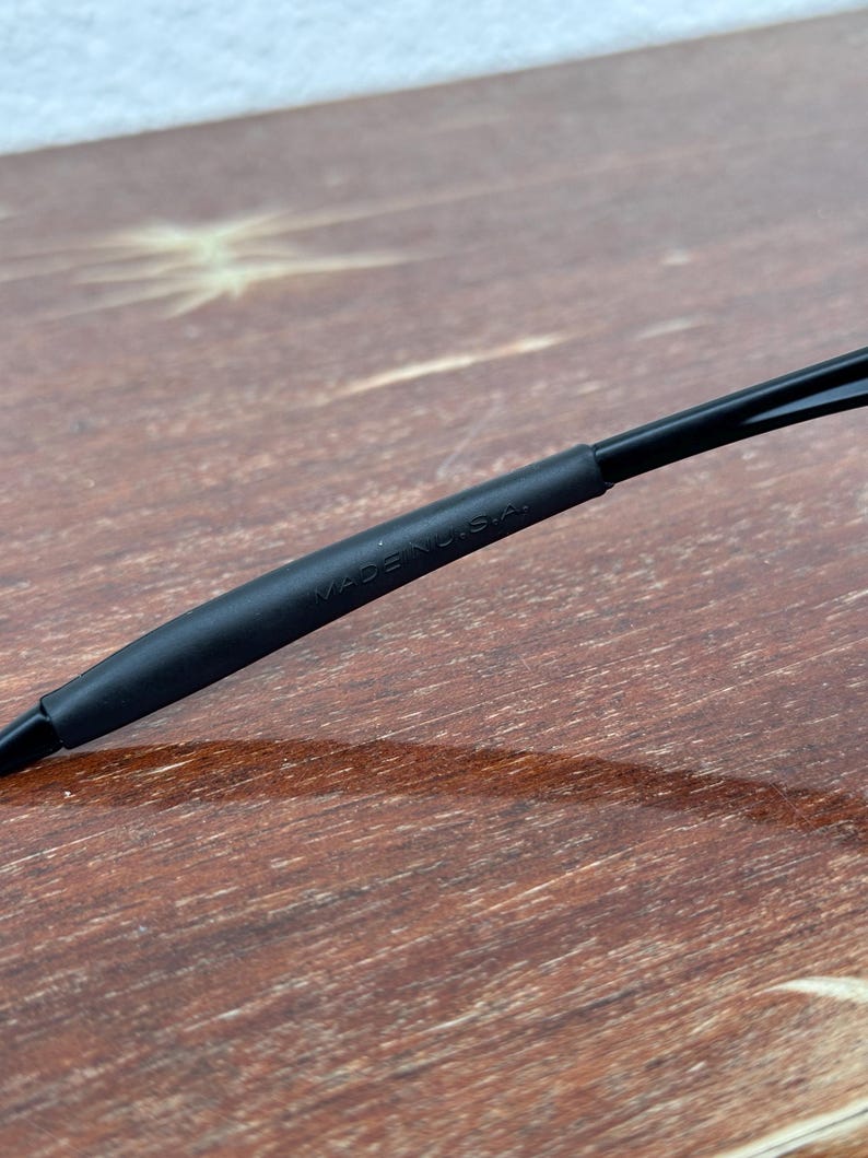 May include: Close-up of a black object with the text "MADE IN U.S.A." printed on it. The object appears to be a part of a pair of glasses or sunglasses. The background is a wooden surface.