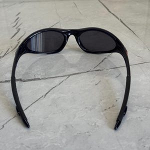 K&ouml;nnte beinhalten: Schwarze Sonnenbrille mit gebogenem Rahmen und dunklen Gl&auml;sern. Die Sonnenbrille hat ein schlankes, sportliches Design, das sich f&uuml;r Outdoor-Aktivit&auml;ten eignet. Die B&uuml;gel der Sonnenbrille sind schwarz und haben eine gummierte Textur.
