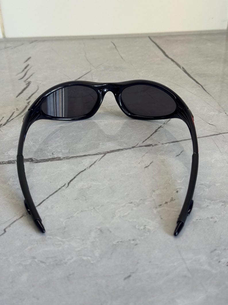 K&ouml;nnte beinhalten: Schwarze Sonnenbrille mit gebogenem Rahmen und dunklen Gl&auml;sern. Die Sonnenbrille hat ein schlankes, sportliches Design, das sich f&uuml;r Outdoor-Aktivit&auml;ten eignet. Die B&uuml;gel der Sonnenbrille sind schwarz und haben eine gummierte Textur.