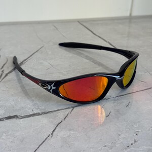 May include: Black sunglasses with red mirrored lenses. The sunglasses have a sleek, sporty design with a star emblem on the side. The frames are black with red accents. The lenses reflect a gradient of red and yellow.