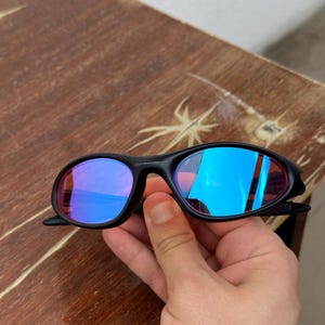May include: Black sunglasses with blue mirrored lenses. The sunglasses have a sleek, sporty design with a curved frame. The lenses reflect a vibrant blue color, and the frame appears to be made of a durable plastic material.