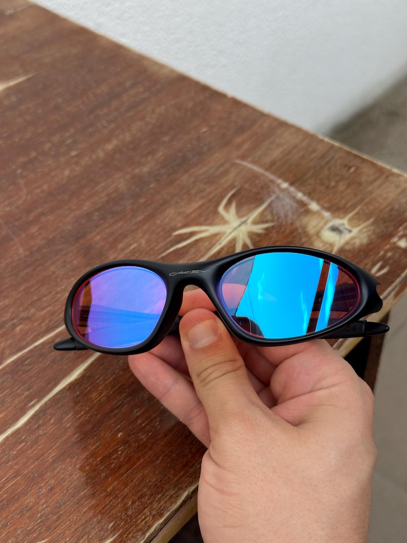 May include: Black sunglasses with blue mirrored lenses. The sunglasses have a sleek, sporty design with a curved frame. The lenses reflect a vibrant blue color, and the frame appears to be made of a durable plastic material.