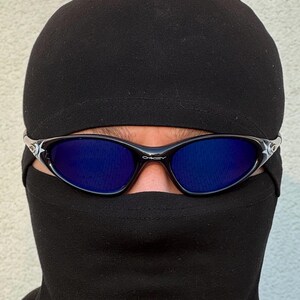 May include: Black sunglasses with blue lenses are worn over a black balaclava. The sunglasses have a black frame with silver accents. The balaclava covers the head and neck, leaving only the eyes visible.