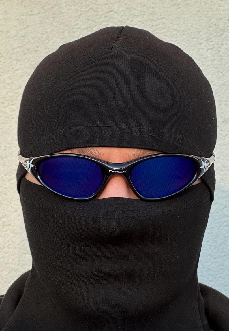 May include: Black sunglasses with blue lenses are worn over a black balaclava. The sunglasses have a black frame with silver accents. The balaclava covers the head and neck, leaving only the eyes visible.