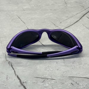 May include: Purple sunglasses with black lenses are displayed against a gray marble surface. The sunglasses have a sleek, modern design with a matte finish. The arms of the sunglasses are black.