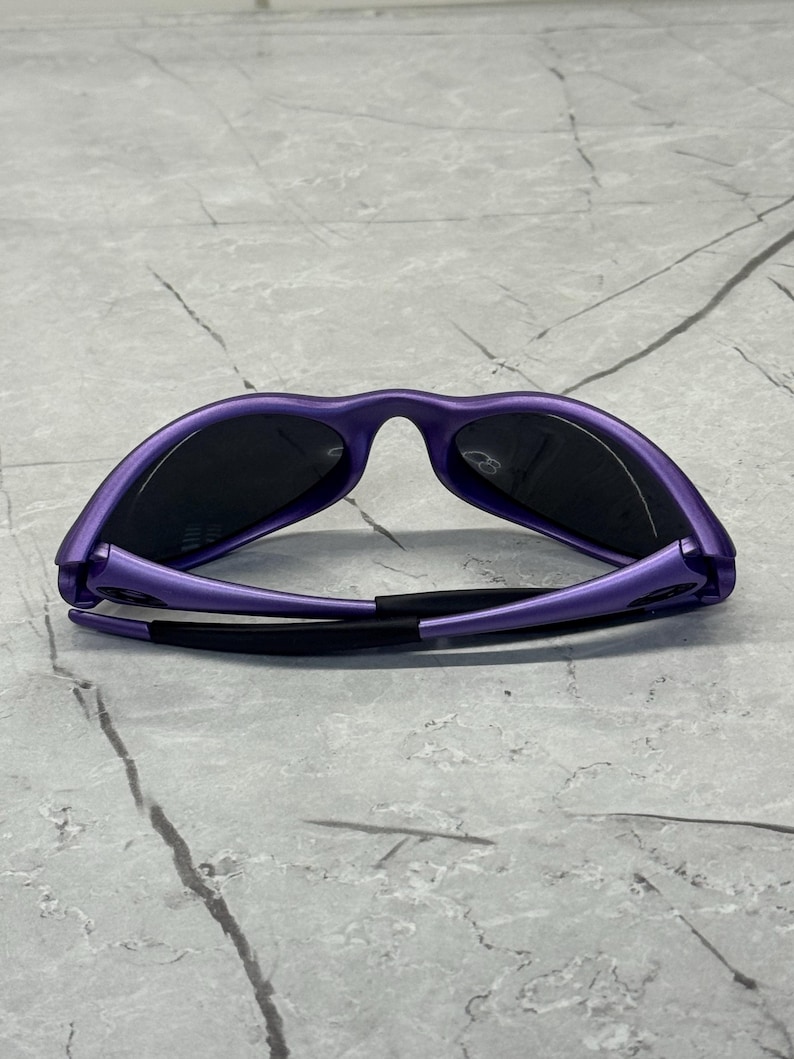 May include: Purple sunglasses with black lenses are displayed against a gray marble surface. The sunglasses have a sleek, modern design with a matte finish. The arms of the sunglasses are black.