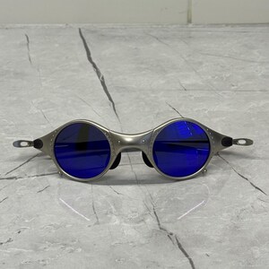 May include: Silver-framed sunglasses with round blue lenses. The sunglasses have a unique, industrial design with visible rivets and a sleek, modern aesthetic. The temples are thin and extend to the ear pieces.