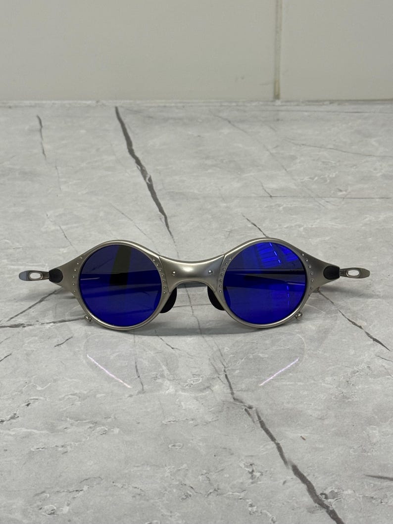 May include: Silver-framed sunglasses with round blue lenses. The sunglasses have a unique, industrial design with visible rivets and a sleek, modern aesthetic. The temples are thin and extend to the ear pieces.