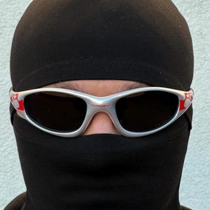 May include: Silver-framed sunglasses with dark lenses and red accents. The sunglasses are worn over a black balaclava. The balaclava covers the head and neck, leaving only the eyes visible.