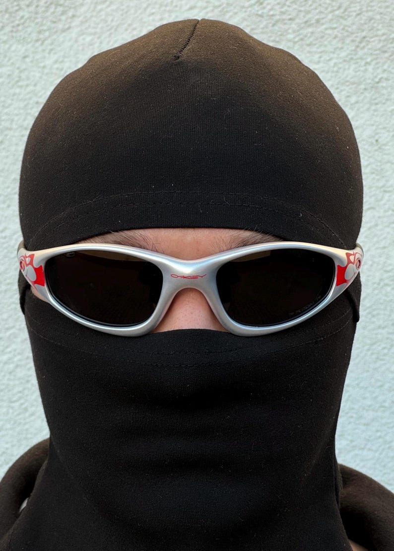 May include: Silver-framed sunglasses with dark lenses and red accents. The sunglasses are worn over a black balaclava. The balaclava covers the head and neck, leaving only the eyes visible.