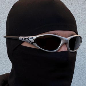 May include: Silver-framed sunglasses with dark lenses are worn over a black balaclava. The sunglasses have a decorative design on the temples. The person is wearing a black hooded sweatshirt.