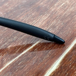May include: Close-up of a black stylus pen with a rounded tip. The pen has a matte finish and a small marking near the tip. The background is a wooden surface with a natural grain.