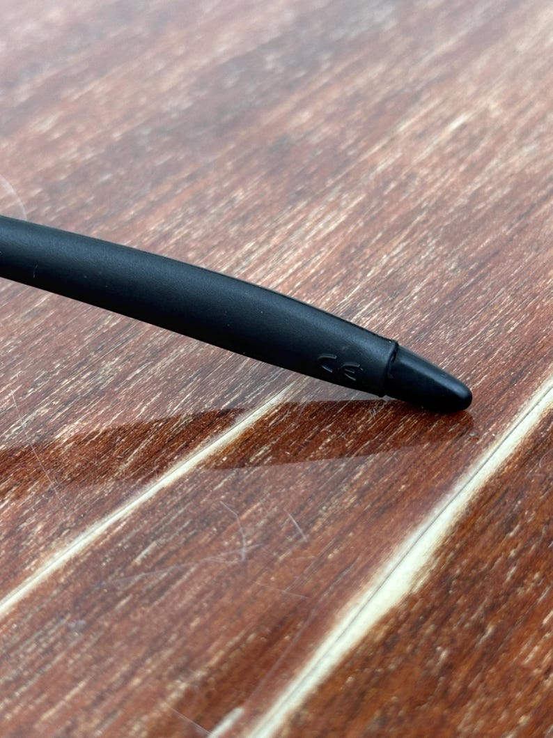 May include: Close-up of a black stylus pen with a rounded tip. The pen has a matte finish and a small marking near the tip. The background is a wooden surface with a natural grain.