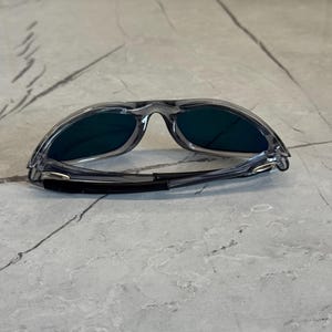 K&ouml;nnte beinhalten: Graue Sonnenbrille mit dunklen Gl&auml;sern. Die Sonnenbrille hat ein sportliches Design mit einem umlaufenden Rahmen und einem schwarzen Band an den B&uuml;geln. Die Gl&auml;ser scheinen eine dunkelgr&uuml;ne oder blaue Farbe zu haben.