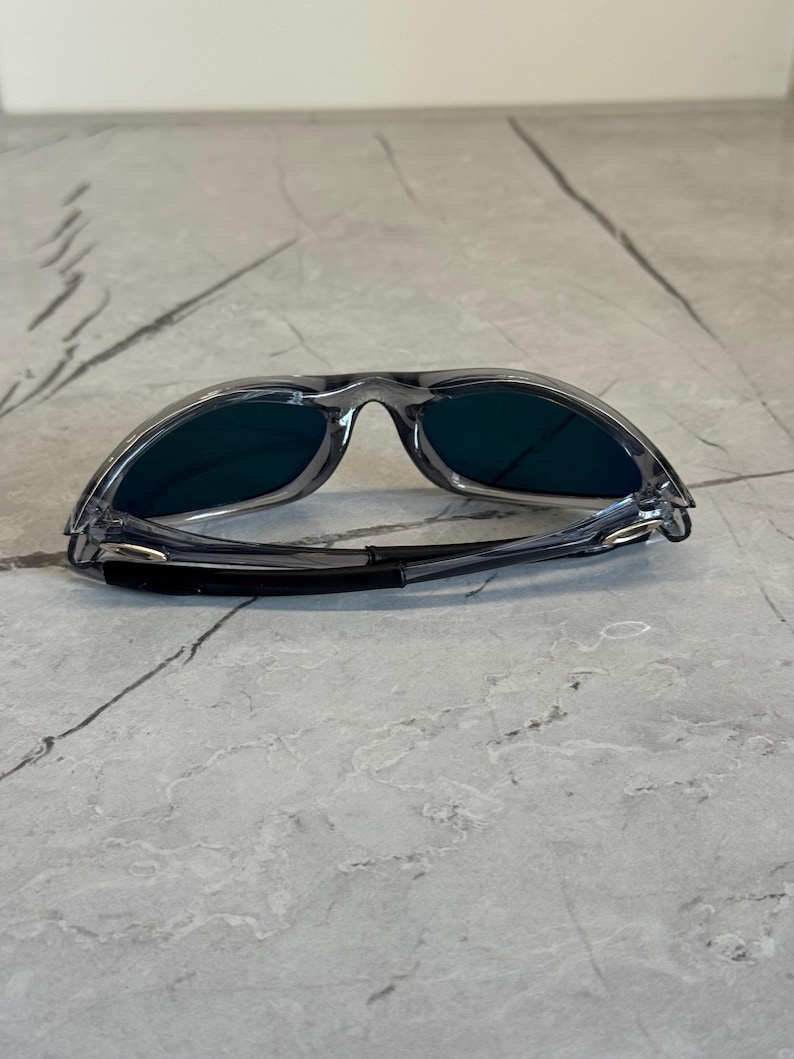 K&ouml;nnte beinhalten: Graue Sonnenbrille mit dunklen Gl&auml;sern. Die Sonnenbrille hat ein sportliches Design mit einem umlaufenden Rahmen und einem schwarzen Band an den B&uuml;geln. Die Gl&auml;ser scheinen eine dunkelgr&uuml;ne oder blaue Farbe zu haben.