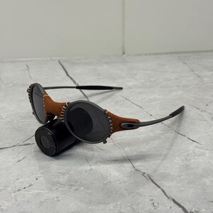 May include: Sunglasses with dark lenses and a brown leather accent stitched to the frame. The sunglasses have a black frame and are resting on a black cylindrical object. The lenses are oval-shaped.