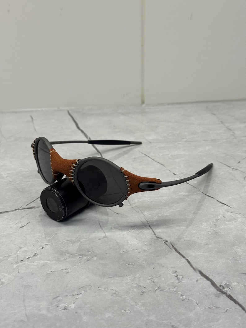 May include: Sunglasses with dark lenses and a brown leather accent stitched to the frame. The sunglasses have a black frame and are resting on a black cylindrical object. The lenses are oval-shaped.