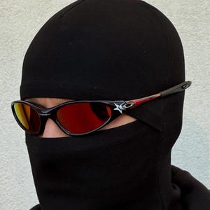 May include: Black sunglasses with red lenses and a black frame. The sunglasses have a white star logo on the side. The person is wearing a black balaclava.