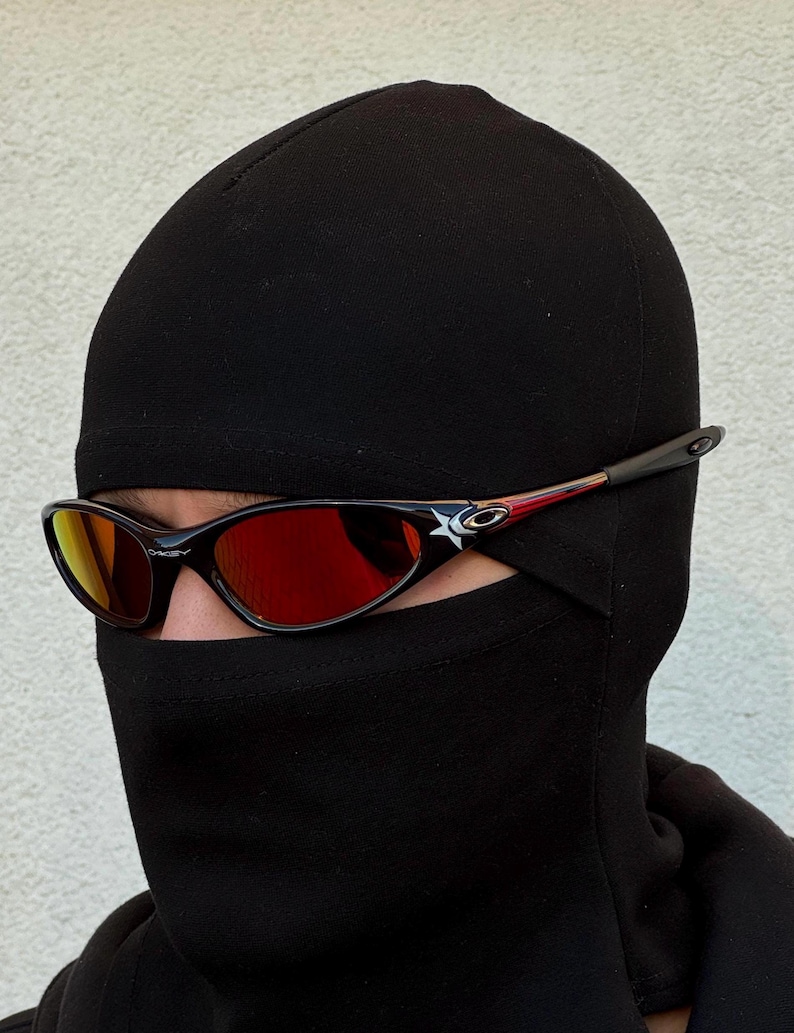 May include: Black sunglasses with red lenses and a black frame. The sunglasses have a white star logo on the side. The person is wearing a black balaclava.