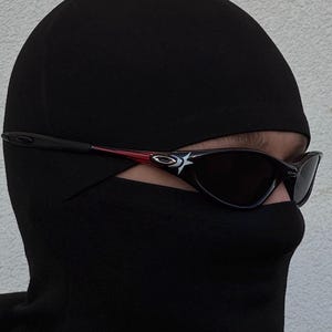 K&ouml;nnte beinhalten: Schwarze Sturmhaube mit dunkler Sonnenbrille. Die Sonnenbrille hat einen schwarzen Rahmen mit roten Akzenten und ein wei&szlig;es Logo an den B&uuml;geln. Die Sturmhaube bedeckt Kopf und Hals, nur die Augen sind sichtbar.