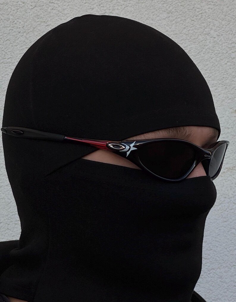 K&ouml;nnte beinhalten: Schwarze Sturmhaube mit dunkler Sonnenbrille. Die Sonnenbrille hat einen schwarzen Rahmen mit roten Akzenten und ein wei&szlig;es Logo an den B&uuml;geln. Die Sturmhaube bedeckt Kopf und Hals, nur die Augen sind sichtbar.