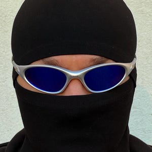 May include: A person wearing a black balaclava and silver-framed sunglasses with blue lenses. The sunglasses are centered on the face, with the black balaclava covering the lower face and head. The background is a plain, light-coloured wall.