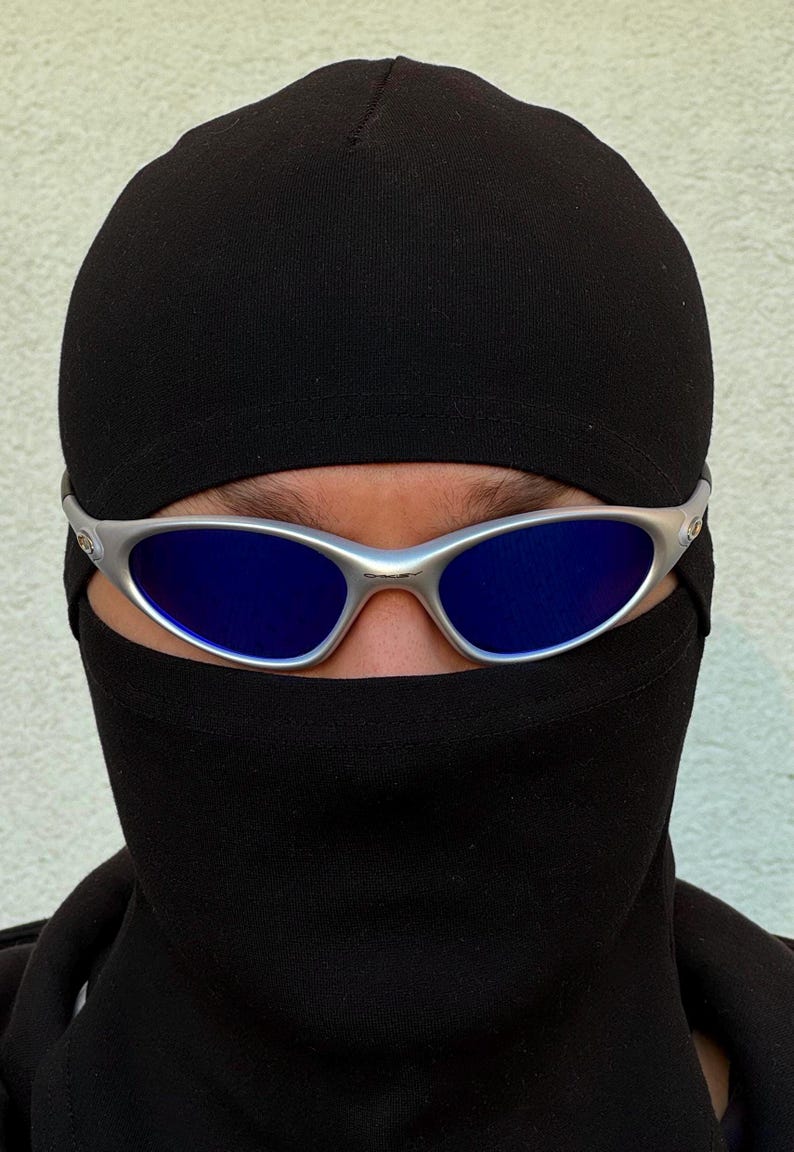 May include: A person wearing a black balaclava and silver-framed sunglasses with blue lenses. The sunglasses are centered on the face, with the black balaclava covering the lower face and head. The background is a plain, light-coloured wall.