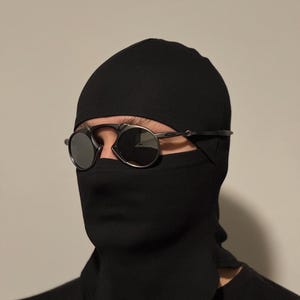 May include: Black balaclava with a face covering and round, dark-tinted sunglasses. The balaclava covers the head and neck, with a slit for the eyes. The sunglasses have a thin, silver frame.