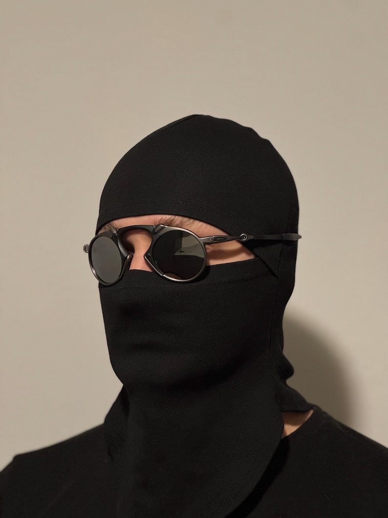 May include: Black balaclava with a face covering and round, dark-tinted sunglasses. The balaclava covers the head and neck, with a slit for the eyes. The sunglasses have a thin, silver frame.