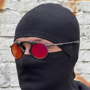 May include: Black balaclava with red-tinted round sunglasses. The sunglasses have a black frame and silver temples. The background is a weathered white brick wall with orange and brown stains.