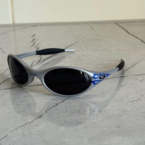 Puede incluir: Gafas de sol plateadas con lentes negras y detalles de llamas azules en las patillas. Las gafas tienen un dise&ntilde;o elegante y moderno, con una montura gris y patillas negras. Las lentes oscuras protegen del sol.