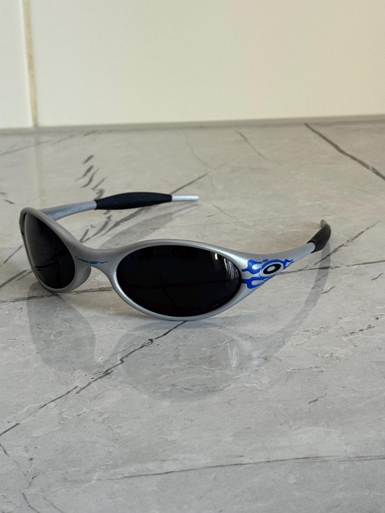 Puede incluir: Gafas de sol plateadas con lentes negras y detalles de llamas azules en las patillas. Las gafas tienen un dise&ntilde;o elegante y moderno, con una montura gris y patillas negras. Las lentes oscuras protegen del sol.