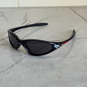 K&ouml;nnte beinhalten: Schwarze Sonnenbrille mit geschwungenem Rahmen und dunklen Gl&auml;sern. Die B&uuml;gel haben ein wei&szlig;es Sterndesign und rote Akzente. Diese Sonnenbrille ist f&uuml;r den Augenschutz und die Mode konzipiert.