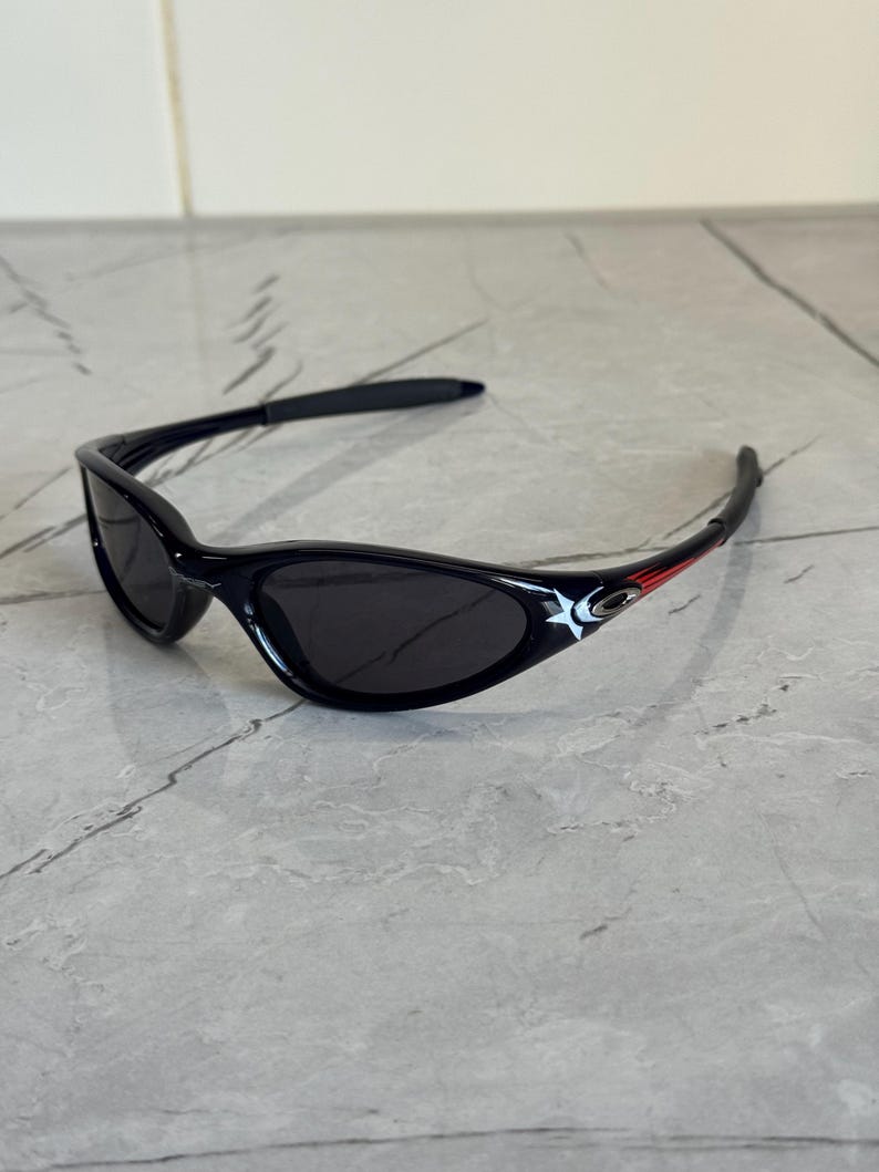 K&ouml;nnte beinhalten: Schwarze Sonnenbrille mit geschwungenem Rahmen und dunklen Gl&auml;sern. Die B&uuml;gel haben ein wei&szlig;es Sterndesign und rote Akzente. Diese Sonnenbrille ist f&uuml;r den Augenschutz und die Mode konzipiert.