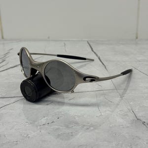 May include: Silver-framed sunglasses with oval lenses and black temple tips. The sunglasses are resting on a black cylindrical object against a gray marble surface. The lenses are a dark gray color.