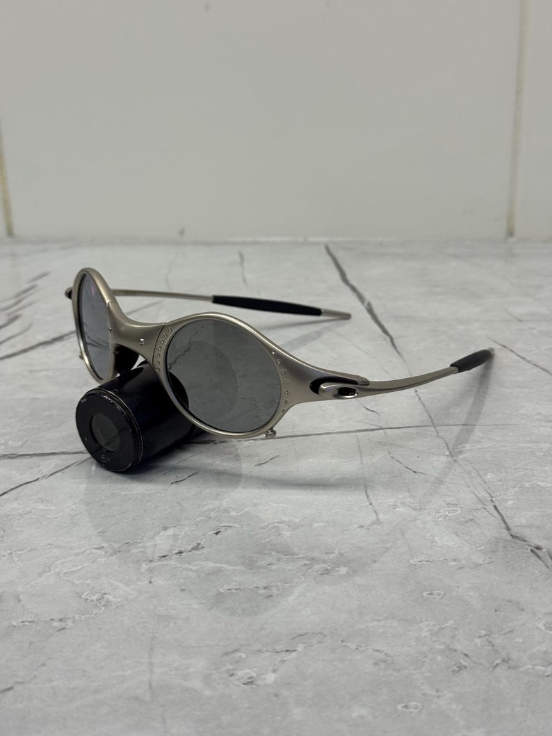 May include: Silver-framed sunglasses with oval lenses and black temple tips. The sunglasses are resting on a black cylindrical object against a gray marble surface. The lenses are a dark gray color.