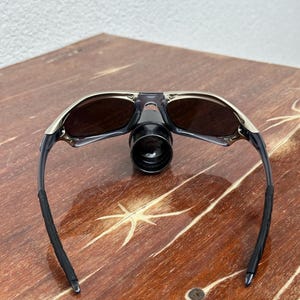 May include: A pair of black and silver sunglasses with dark lenses. The sunglasses are folded in half and are sitting on a wooden surface.