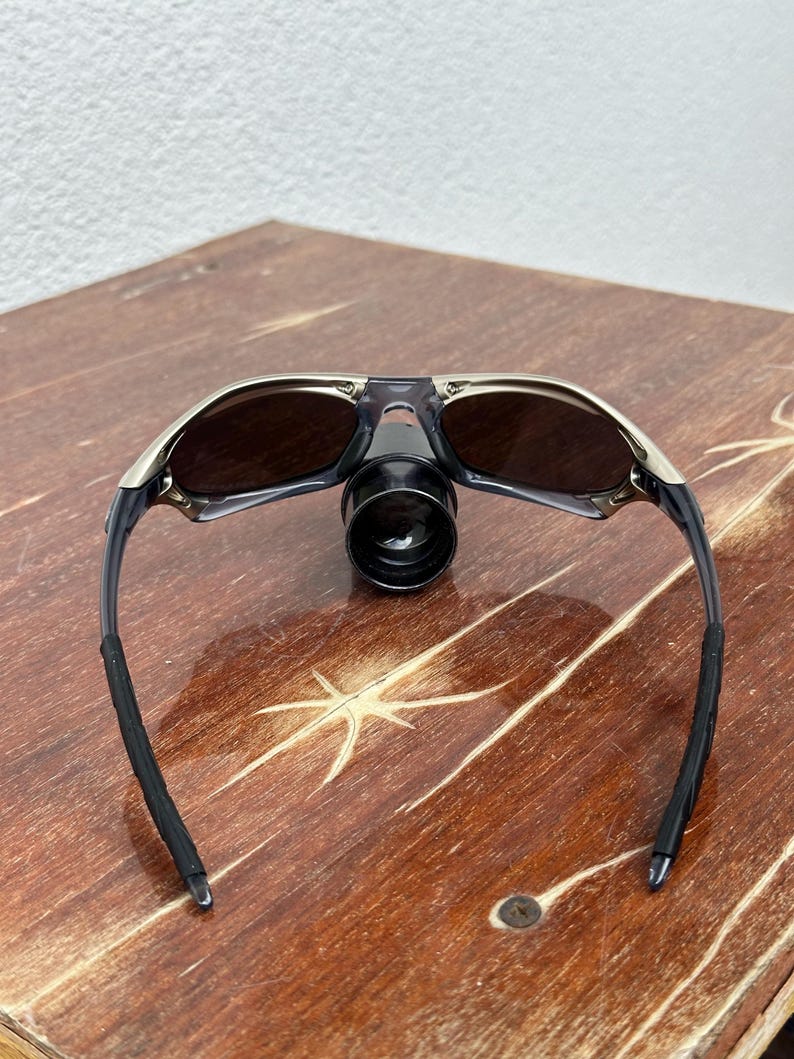 May include: A pair of black and silver sunglasses with dark lenses. The sunglasses are folded in half and are sitting on a wooden surface.