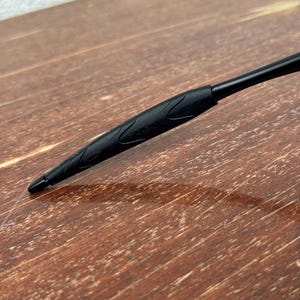 Op de afbeelding: Close-up van een zwarte pen met een getextureerde grip en een puntige punt. De pen staat schuin over een houten oppervlak, wat het strakke ontwerp en de functionaliteit laat zien. De grip van de pen heeft een subtiel, decoratief patroon.