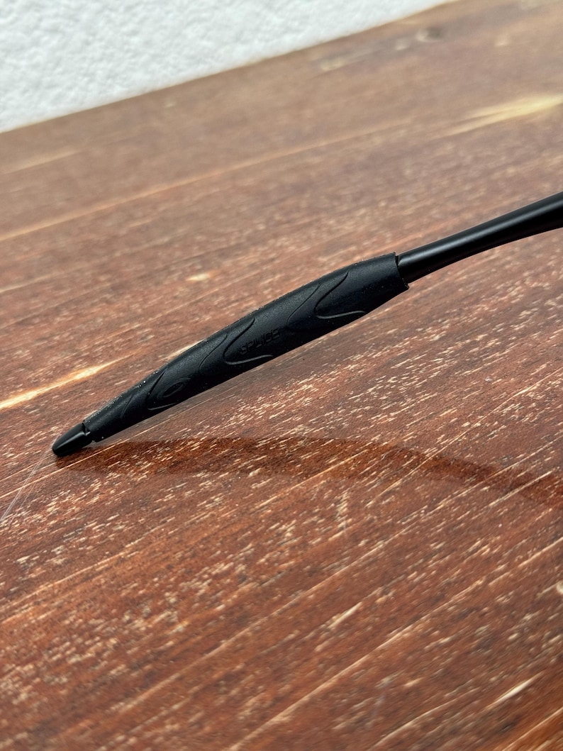 Op de afbeelding: Close-up van een zwarte pen met een getextureerde grip en een puntige punt. De pen staat schuin over een houten oppervlak, wat het strakke ontwerp en de functionaliteit laat zien. De grip van de pen heeft een subtiel, decoratief patroon.