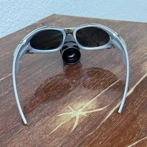 Puede incluir: Gafas de seguridad grises y negras con un accesorio cil&iacute;ndrico negro. Las gafas tienen lentes oscuras y una montura gris claro. Las gafas est&aacute;n sobre una superficie de madera.