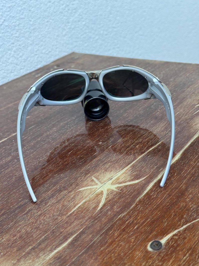 Puede incluir: Gafas de seguridad grises y negras con un accesorio cil&iacute;ndrico negro. Las gafas tienen lentes oscuras y una montura gris claro. Las gafas est&aacute;n sobre una superficie de madera.