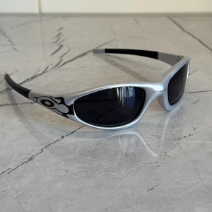 May include: Silver-framed sunglasses with black lenses. The sunglasses have a curved design with black accents on the temples and arms. The lenses are dark, and the frame has a sleek, modern look. The sunglasses are designed for eye protection.