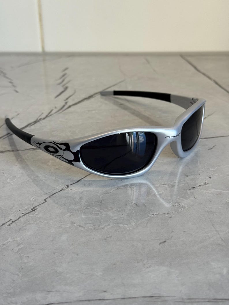 May include: Silver-framed sunglasses with black lenses. The sunglasses have a curved design with black accents on the temples and arms. The lenses are dark, and the frame has a sleek, modern look. The sunglasses are designed for eye protection.