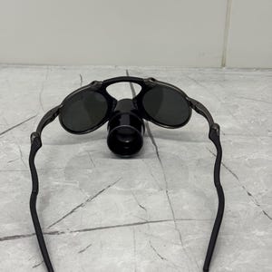 以下が含まれることがあります： 楕円形のレンズと中央の円筒形のアタッチメントが付いた黒いサングラス。フレームは暗く、レンズは濃い緑色です。サングラスのアームはレンズから離れるように湾曲しています。