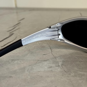 May include: Close-up of silver sunglasses with black lenses and arms. The word "straight" is printed on the silver frame. The sunglasses are on a light gray surface.