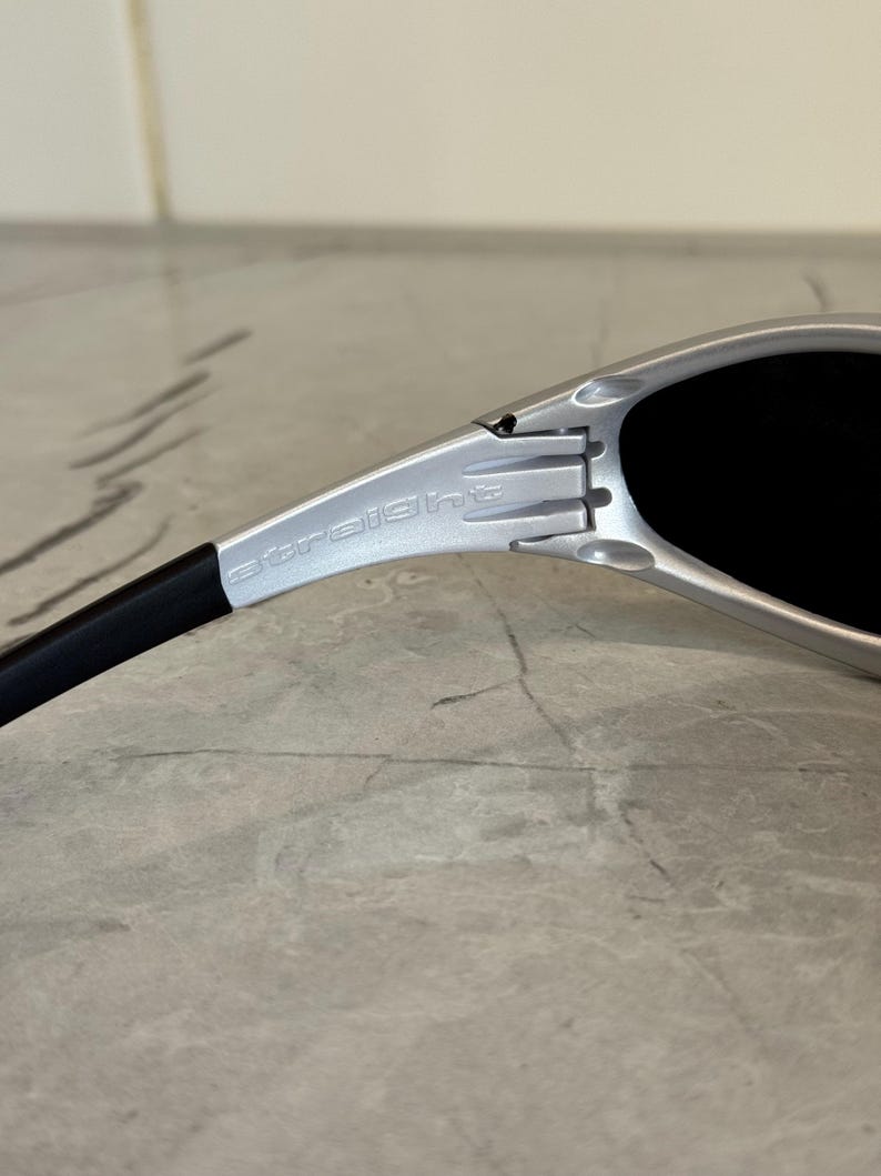 May include: Close-up of silver sunglasses with black lenses and arms. The word "straight" is printed on the silver frame. The sunglasses are on a light gray surface.
