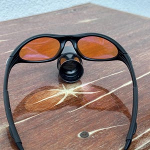 May include: Black sports sunglasses with orange-tinted lenses. The sunglasses have a sleek, curved design and a small, cylindrical attachment between the lenses. The frame is black, and the lenses are a vibrant orange color. The sunglasses are resting on a wooden surface.