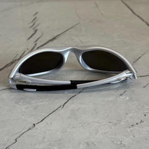 May include: Silver sunglasses with dark lenses. The sunglasses have a sleek, modern design with a curved frame and a black band at the temples. The lenses are dark, providing shade and protection from the sun.